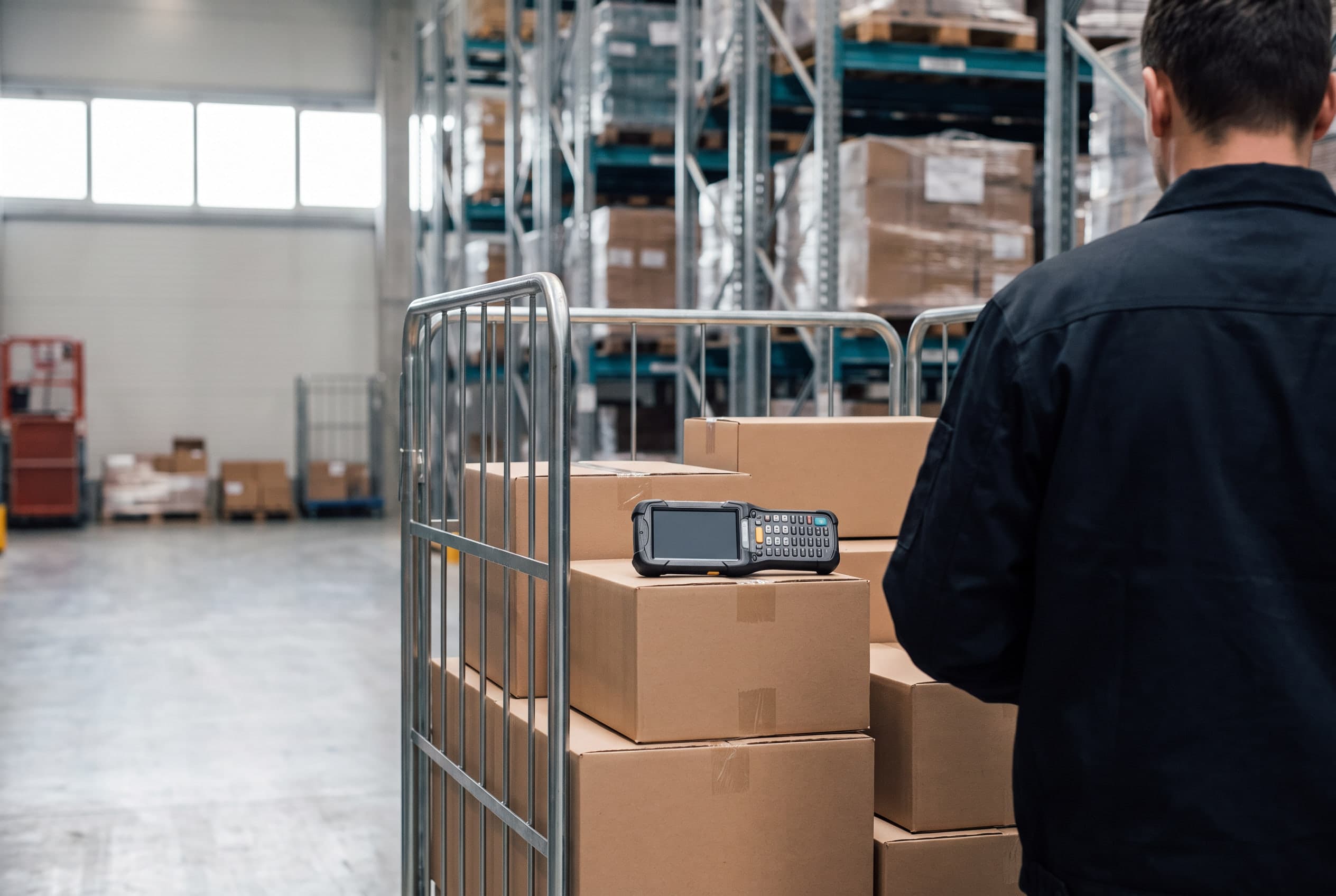 Orderpickplek in een magazijn: een rolcontainer met ingepakte dozen en een handscanner, stellingen op de achtergrond.